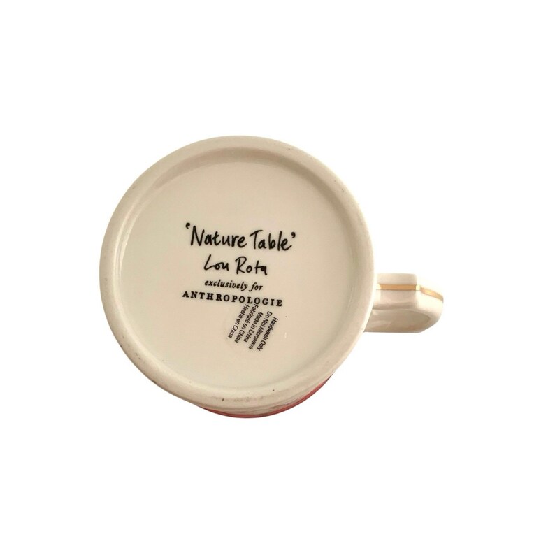 May include: A white ceramic mug with a red stripe at the bottom and a gold-colored handle. The mug's interior features the text "Nature Table" by Lou Rota, exclusively for Anthropologie.