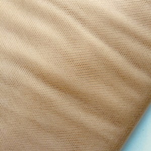 Può includere: Un primo piano di un tessuto di tulle beige con una trama a rete fine. Il tessuto è piegato e ha un aspetto morbido e delicato.