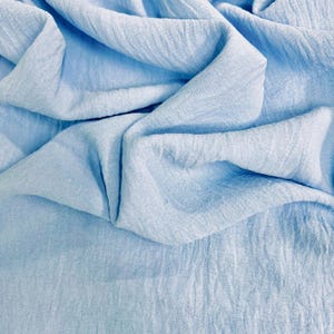 May include: Close-up of a light blue fabric with a crinkled texture. The fabric is draped and folded, creating shadows and highlights. The material appears soft and lightweight, suitable for clothing or home decor.