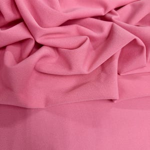 Puede incluir: Primer plano de una tela rosa con pliegues suaves y una superficie texturizada. La tela es de un color rosa sólido y vibrante, con sutiles variaciones de tono debido a los pliegues y las sombras. El material parece ser un textil suave y drapeado.