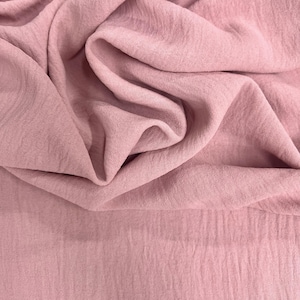 May include: Close-up of a dusty rose fabric with a crinkled texture. The fabric is draped and folded, creating shadows and highlights. The material appears soft and lightweight, suitable for clothing or home decor.