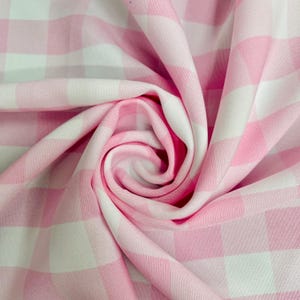 May include: Close-up of a pink and white gingham fabric. The fabric is folded and draped, showcasing the pattern's alternating squares. The material appears soft and lightweight, suitable for various sewing projects.
