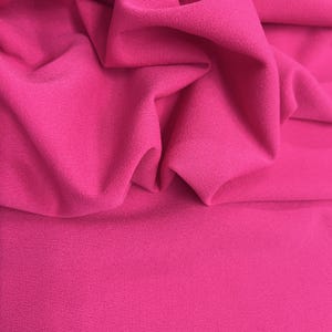 Puede incluir: Primer plano de una vibrante tela fucsia con una superficie texturizada. La tela está doblada y drapeada, mostrando su textura suave y su rico color. Ideal para ropa, decoración del hogar o proyectos de manualidades.