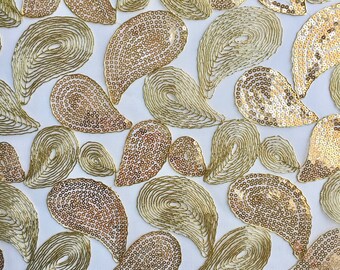 Może przedstawiać: Biały materiał z złotym wzorem paisley. Kształty paisley są wykonane z cekinów i obwiedzione złotą nicią.
