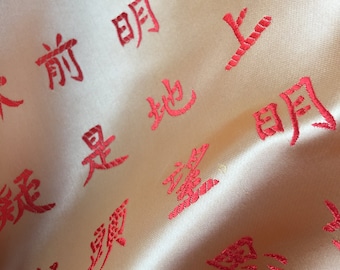 Könnte beinhalten: Nahaufnahme eines beigefarbenen Stoffes mit roten, gestickten chinesischen Schriftzeichen. Die Schriftzeichen sind in verschiedenen Größen und Stilen gehalten und in einem zufälligen Muster angeordnet. Der Stoff ist weich und glatt und hat einen dezenten Glanz.