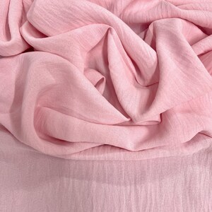 May include: A close-up shot of a soft, flowing fabric in a delicate shade of pink. The fabric is draped and folded, creating a sense of texture and movement. The material appears lightweight and possibly sheer, with subtle variations in color.