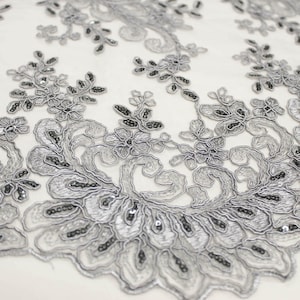 May include: Silver and black floral lace fabric with a delicate pattern. The lace has a beaded design and is perfect for adding a touch of elegance to any project.