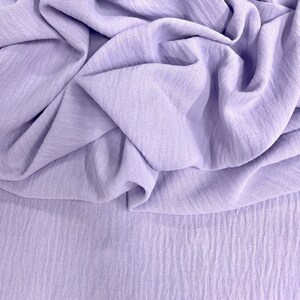May include: A close-up shot of a light purple fabric with a crinkled texture. The fabric is draped and folded, creating shadows and highlights. The material appears soft and lightweight, suitable for clothing or home decor.