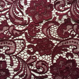 May include: Burgundy lace fabric with a delicate floral pattern. The lace features intricate swirls and a repeating design of small flowers.