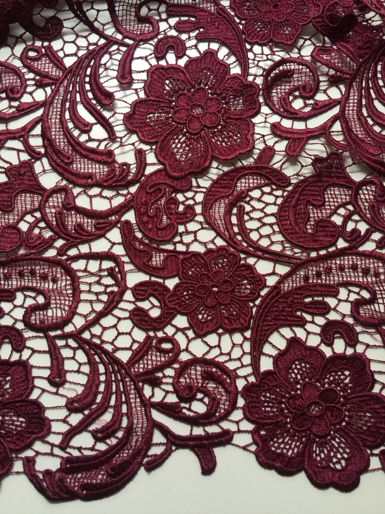 May include: Burgundy lace fabric with a delicate floral pattern. The lace features intricate swirls and a repeating design of small flowers.