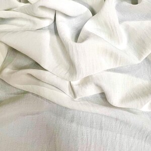 May include: Close-up of a white fabric with soft folds and a slightly textured surface. The fabric appears to be lightweight and flowing, with subtle variations in tone and shadow, creating a sense of depth and movement. Ideal for apparel or home decor.