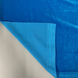 May include: Close-up of a turquoise blue velvet fabric with a turquoise blue lining. The fabric is folded over to show the lining. A ruler is visible at the bottom of the image with measurements in inches.