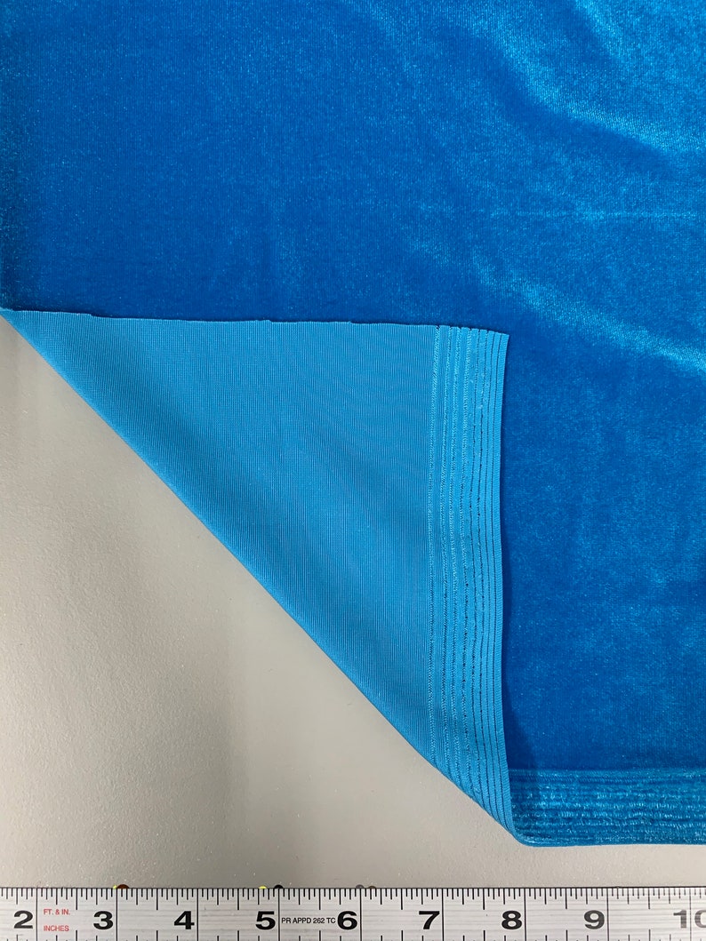 May include: Close-up of a turquoise blue velvet fabric with a turquoise blue lining. The fabric is folded over to show the lining. A ruler is visible at the bottom of the image with measurements in inches.