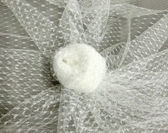 May include: White tulle fabric with a delicate dotted pattern. The fabric is draped and gathered, with a rolled bundle of the same material in the center. The overall effect is soft and airy, suitable for bridal or craft projects.