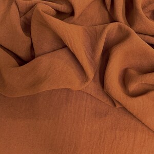 May include: Close-up of a flowing, textured fabric in a warm, earthy orange-brown hue. The material is draped and folded, creating soft shadows and highlights. The fabric appears to be lightweight and suitable for clothing or home decor.