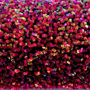 May include: A close-up of a fabric covered in iridescent sequins. The sequins are a mix of pink, red, and gold colors. The fabric is a dark black color.
