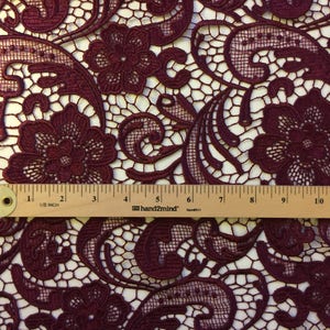 May include: Burgundy lace fabric with a floral pattern. The fabric is measured with a ruler that shows 12 inches.