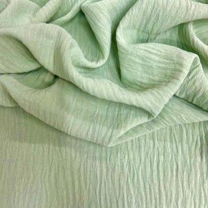 May include: A close-up shot of a light green fabric with a textured, crinkled appearance. The fabric is draped in soft folds, creating shadows and highlights. The material appears lightweight and flowing, suitable for clothing or home decor.