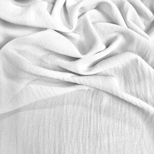 May include: A close-up shot of white fabric with soft folds and a textured surface. The fabric appears to be lightweight and flowing, with subtle variations in tone and shadow, creating a sense of depth and movement.