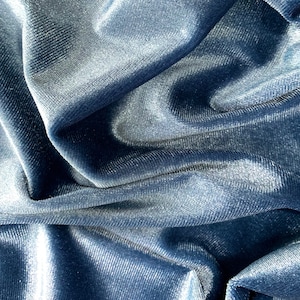May include: Close-up of a soft, blue velvet fabric with a subtle sheen. The fabric is draped and folded, creating a textured and luxurious look.