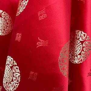 May include: Red fabric with a gold embroidered pattern. The pattern includes a repeating design of a circle with a stylized flower inside and the Chinese character for "fortune" written in gold.