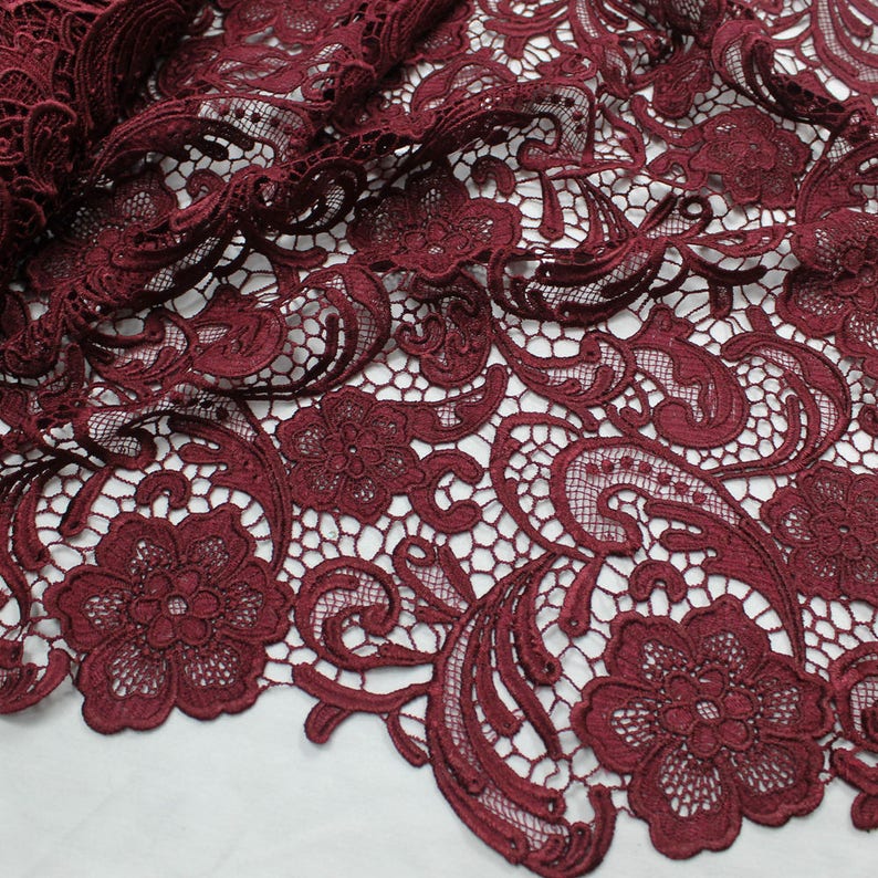 May include: Burgundy lace fabric with a delicate floral pattern. The lace features intricate swirls and paisley designs.