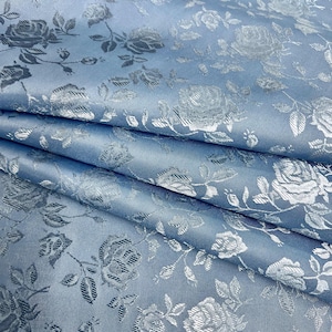 May include: A folded, light blue fabric with a repeating pattern of silver roses and leaves. The fabric has a subtle sheen, and the floral design is intricately detailed, creating a luxurious appearance. The folds add depth and texture to the textile.