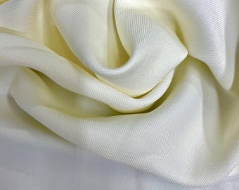 Puede incluir: Primer plano de una tela plegada de color blanquecino con un brillo sutil. La tela tiene una textura suave y fluida con líneas de tejido diagonales visibles. El color pasa de un amarillo pálido a un blanco cremoso.