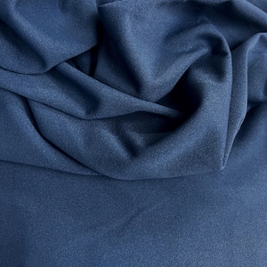 Op de afbeelding: Close-up van een donkerblauwe stof met zachte plooien en een matte afwerking. De stof is effen van kleur, met subtiele tintvariaties door de draperie. Het materiaal wordt waarschijnlijk gebruikt voor kleding of interieurdecoratie.