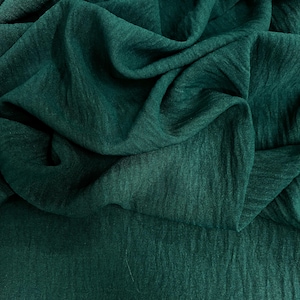 May include: Close-up of a dark teal fabric with a textured, slightly wrinkled appearance. The fabric is draped and folded, creating shadows and highlights that emphasize its texture. The overall effect is of a soft, flowing material.