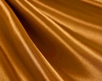 Op de afbeelding: Een close-up van een gladde, goud-bruine stof met een subtiele glans. De stof is gedrapeerd en gevouwen, waardoor zachte, vloeiende lijnen ontstaan.