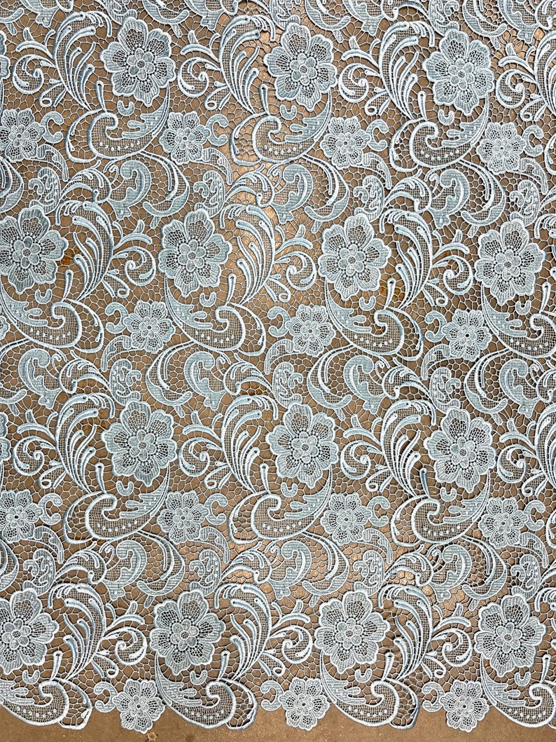 May include: A delicate white lace fabric with a floral pattern in a light blue color. The lace has a delicate, intricate design with small flowers and leaves.