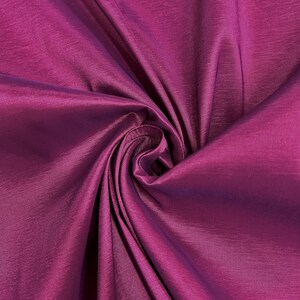 Puede incluir: Primer plano de una tela fucsia vibrante con un brillo lustroso. El material está recogido y doblado, creando un patrón arremolinado de luz y sombra. La textura de la tela parece suave y fluida, lo que sugiere que es un textil lujoso.