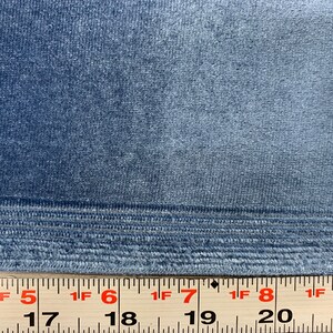 May include: Close-up of a blue velvet fabric with a subtle sheen. The fabric has a textured surface and a woven edge. A ruler is visible at the bottom of the image, with markings in inches.
