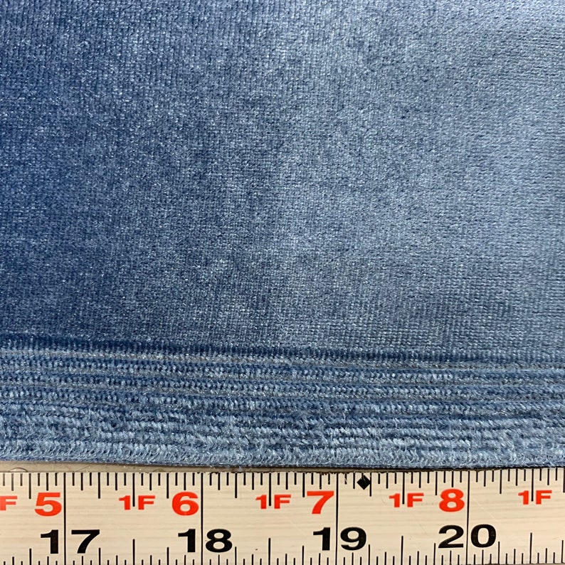 May include: Close-up of a blue velvet fabric with a subtle sheen. The fabric has a textured surface and a woven edge. A ruler is visible at the bottom of the image, with markings in inches.