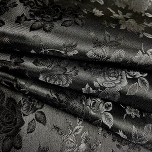 May include: Close-up of black fabric with a floral rose pattern. The fabric is folded, showing the texture and sheen of the material. The roses and leaves are subtly raised, creating a textured effect.