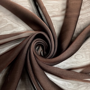 May include: Close-up of a swirl of brown sheer fabric. The fabric is a rich, dark brown color and appears to be lightweight and flowing. The fabric is arranged in a spiral, creating a sense of depth and texture. The background is a light wood grain.