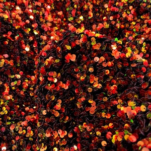 Può includere: Primo piano di un tessuto con sfondo nero ricoperto di paillettes iridescenti in tonalità di rosso, arancione e oro.