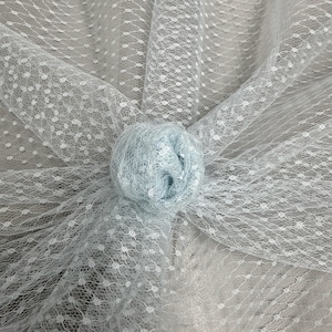 May include: A close-up of light blue tulle fabric with a delicate dotted pattern. A gathered, round, light blue fabric flower is centered on the tulle. The fabric is soft and airy, suitable for crafting or decorative purposes.