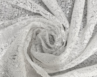 May include: A close-up shot of a white, shimmering fabric covered in small, round sequins. The fabric is draped and swirled, creating a textured, reflective surface. The sequins catch the light, giving the fabric a sparkling appearance. Ideal for apparel and decor.