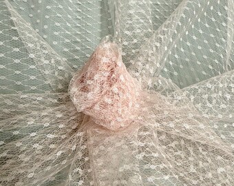 May include: Close-up of delicate, off-white lace fabric with a repeating dot pattern. A gathered, textured, light pink fabric is nestled within the lace, creating a layered effect. The background is a soft, muted green.