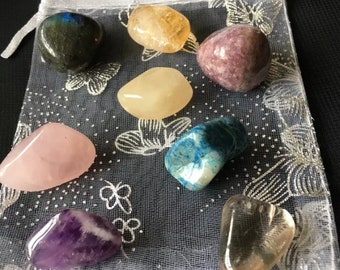 May include: A collection of polished gemstones in various colors, including pink, purple, yellow, and blue, displayed on a sheer, floral-patterned pouch. The stones vary in size and shape, offering a diverse assortment.