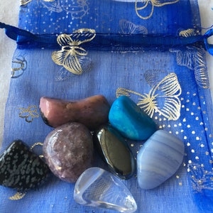 May include: A selection of polished gemstones in shades of pink, blue, and black, presented in a sheer blue drawstring bag adorned with gold butterfly patterns. The bag features a tag with the text "Earthstarsbygail".