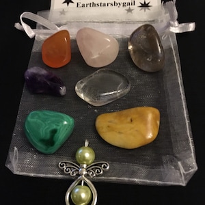 Op de afbeelding: Een verzameling gepolijste edelstenen in verschillende kleuren, waaronder oranje, roze, paars, helder, groen en geel, gepresenteerd in een doorzichtige witte zak met trekkoord. Een zilveren engel hanger met groene parel accenten is ook inbegrepen. De zak heeft een label met "earthstarsbygail."