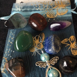 Può includere: Una collezione di pietre preziose lucide in vari colori, tra cui verde, viola e blu, presentate in un sacchetto di organza blu trasparente. È incluso anche un ciondolo a forma di angelo argentato con perline blu. La borsa ha il testo "Earthstarsbygail".