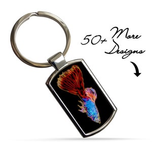 May include: A silver keychain with a black rectangular tag featuring a neon-colored guppy fish with red, orange, blue, and yellow accents. The text "50+ More Designs" is visible in the background.