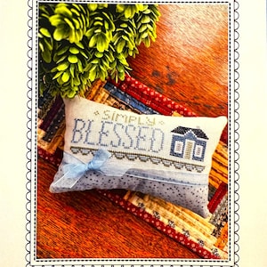 May include: A decorative pillow with the text "Simply Blessed" embroidered in blue thread, featuring a small house design. The pillow is adorned with a light blue ribbon and rests on a patchwork quilt. The image is framed with a decorative border.