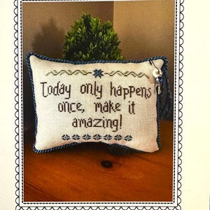 May include: A decorative pillow with the embroidered text "Today only happens once, make it amazing!". The pillow has a blue border and is decorated with floral patterns. A small key charm hangs from the side. The image is framed with a scalloped border.