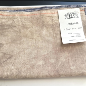 May include: A piece of beige linen fabric with a blue stitched border and an orange stitch detail. A white tag reads "AT RANCH FABRICS" and "Mirkwood". The tag also lists "Aida", "Linen 32", and measurements of 17x28, 26x36, and 35x52.