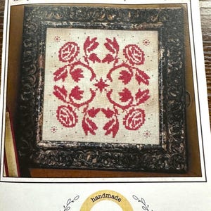 May include: A framed reproduction pattern of an antique coverlet pillow. The design features a red floral pattern on a beige background. The frame is dark brown with ornate detailing. The text "Red Haven #5 Rose Coverlet" is at the top.